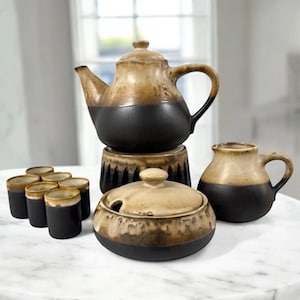 May include: A ceramic tea set featuring a teapot on a warmer, a lidded sugar bowl, a creamer, and six small cups. Each piece has a two-tone design with a matte black base and a glazed tan top, showcasing a drip-style transition between colors.