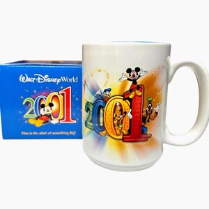May include: A white ceramic mug with a large handle, featuring the characters Mickey Mouse, Donald Duck, Goofy, and Pluto. The mug has the year "2001" in colorful lettering. A blue box with the words "Walt Disney World" and "2001" is next to the mug.
