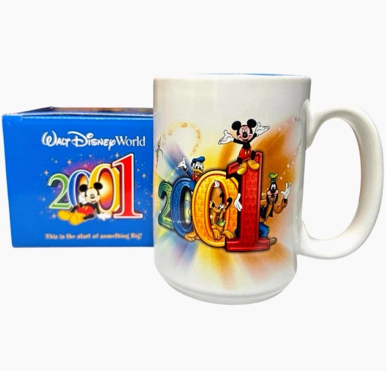 May include: A white ceramic mug with a large handle, featuring the characters Mickey Mouse, Donald Duck, Goofy, and Pluto. The mug has the year "2001" in colorful lettering. A blue box with the words "Walt Disney World" and "2001" is next to the mug.