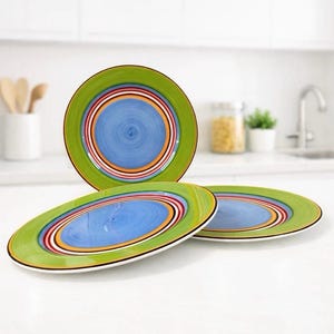 Puede incluir: Tres platos redondos de cerámica con un patrón circular concéntrico. Cada plato tiene un centro azul brillante rodeado por anillos de color naranja, blanco y rojo oscuro, con un borde ancho de color verde lima en el exterior.