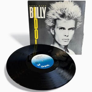 Puede incluir: Un disco de vinilo y la portada del álbum "Don't Stop" de Billy Idol. La portada del álbum presenta un retrato en blanco y negro de Billy Idol con el título del álbum en texto amarillo y blanco. El disco es negro con una etiqueta azul y blanca.