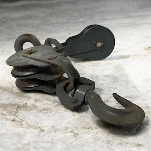May include: A weathered metal pulley system, featuring a double-wheel design and a large hook. The pulley is painted grey and shows signs of use. The hook is curved and robust, indicating its industrial purpose. The item appears to be vintage.