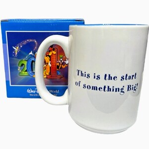 May include: White ceramic mug with the text "This is the start of something Big!" in blue. The mug features a handle and a colorful image of Disney characters. A blue box with Disney World graphics is behind the mug.