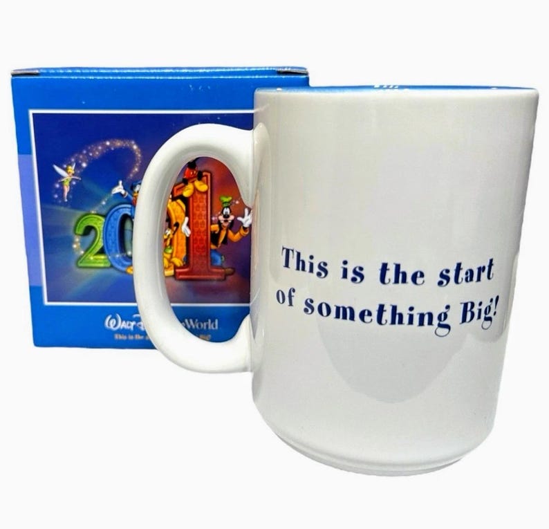 May include: White ceramic mug with the text "This is the start of something Big!" in blue. The mug features a handle and a colorful image of Disney characters. A blue box with Disney World graphics is behind the mug.