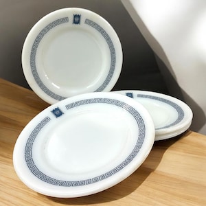 May include: A collection of white ceramic plates with a blue Greek key border design. Each plate has a small blue emblem. The plates are displayed on a wooden surface, highlighting their clean, classic aesthetic. A classic addition to any kitchen.