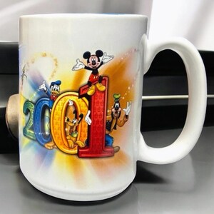 May include: White ceramic mug featuring the characters Mickey Mouse, Donald Duck, Pluto, and Goofy. The mug has the number "2001" in colorful letters and numbers. The mug has a white handle and a blue interior.