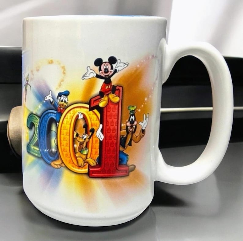 May include: White ceramic mug featuring the characters Mickey Mouse, Donald Duck, Pluto, and Goofy. The mug has the number "2001" in colorful letters and numbers. The mug has a white handle and a blue interior.