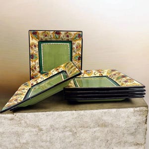May include: A set of square ceramic plates featuring a solid sage green center bordered by a decorative rim with a repeating floral pattern in shades of red, yellow, and green. The plates have a dark brown edge and are stacked together.