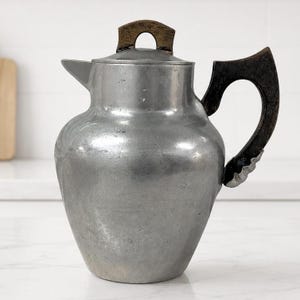 May include: A metallic pitcher with a matte gray finish, featuring a rounded body, a pointed spout, and a lid with a dark brown wooden handle. A matching dark brown wooden handle is attached to the side, secured with a decorative metal accent.