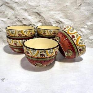 May include: A set of four ceramic bowls featuring a cream-colored interior and a decorative exterior band. The pattern includes yellow arches, green stripes, and red floral motifs with black accents. The bowls are arranged in a small stack.
