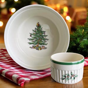 Puede incluir: Un cuenco de cerámica blanco y un pequeño ramequín con diseños de árbol de Navidad y acebo. El cuenco presenta una detallada ilustración de un árbol de Navidad verde. El ramequín tiene un borde verde y un diseño de ramita de acebo.