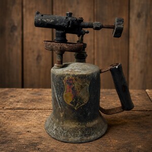 May include: A vintage metal blowtorch featuring a weathered, dark gray finish with visible rust on the handle and burner assembly. The cylindrical base displays a faded, shield-shaped emblem in yellow, red, and black, with a black handle attached.