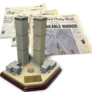 Könnte beinhalten: Ein detailliertes Modell der Twin Towers mit umliegenden Gebäuden, auf einem dekorativen Sockel. Das Modell wird von Zeitungsausschnitten mit Schlagzeilen über die Anschläge vom 11. September 2001 begleitet. Eine Plakette mit einem Zitat ist ebenfalls enthalten.