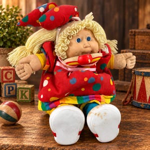Pode incluir: Uma boneca com cabelo de lã loiro e olhos azuis vestindo um vestido vermelho com bolinhas multicoloridas e um laço combinando. A boneca tem um corpo de tecido macio, rosto de plástico e sapatos brancos. Possui gola e bainha listradas.