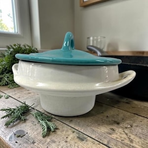 May include: A cream-colored ceramic casserole dish with a turquoise lid. The dish has a wide, flared base and two small handles. The lid has a loop handle. The dish is on a wooden surface.