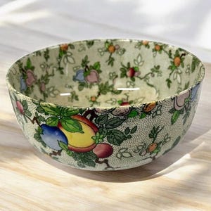 May include: A round ceramic bowl featuring a cream-colored base with a dense, stippled texture. The exterior and interior are decorated with a repeating pattern of colorful fruit, including yellow peaches, blue plums, and green leaves on brown vines.