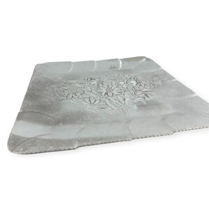 Może przedstawiać: Srebrna metalowa taca z kwiatowym wzorem. Taca ma prostokątny kształt z falistą krawędzią i szczeg&oacute;łowym wytłoczonym wzorem kwiatowym pośrodku. Taca jest lekko pochylona.