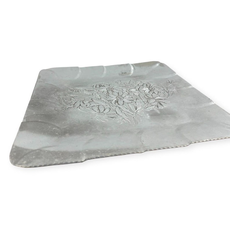 Może przedstawiać: Srebrna metalowa taca z kwiatowym wzorem. Taca ma prostokątny kształt z falistą krawędzią i szczeg&oacute;łowym wytłoczonym wzorem kwiatowym pośrodku. Taca jest lekko pochylona.