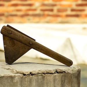 May include: A vintage hand saw with a wooden handle and a rusty, triangular blade. The saw is resting on a concrete surface, with a blurred brick wall in the background. The saw's design suggests it is an older tool.