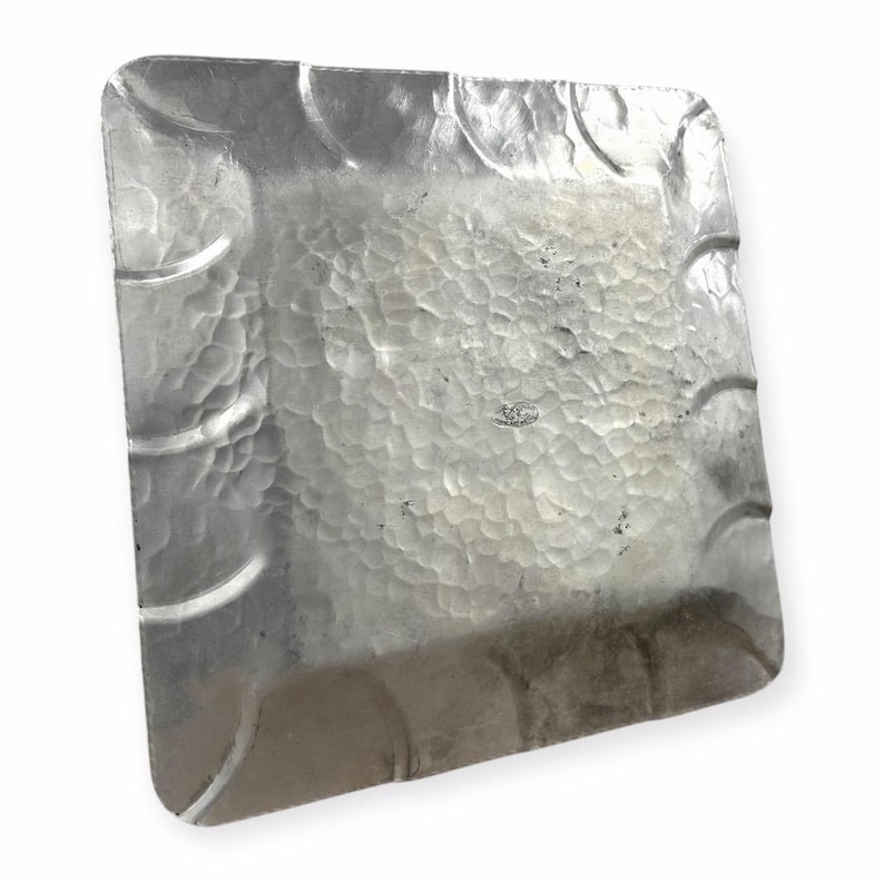 Może przedstawiać: Kwadratowa, srebrna metalowa taca o młotkowanej fakturze. Taca ma zaokrąglone rogi i dekoracyjne zakrzywione akcenty wzdłuż krawędzi. Powierzchnia ma wygląd z wgłębieniami, a og&oacute;lny design jest prosty i elegancki.
