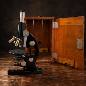 May include: A black metal compound microscope with a silver eyepiece tube and multiple brass objective lenses, positioned next to an open wooden storage case featuring a metal latch and a small rectangular label on the interior door panel.