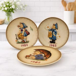 May include: Three decorative plates with vintage illustrations. One plate features a girl with a basket, another a mail carrier, and the third a child under an umbrella. Each plate has a year printed on it: 1984, 1983, and 1982. The plates have a cream-colored background.