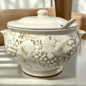 Puede incluir: Sopera de cerámica color crema con tapa y cuchara pequeña. La sopera presenta un diseño en relieve de racimos de uvas y hojas. Las asas son pequeñas y curvas. La sopera está sobre una superficie blanca.