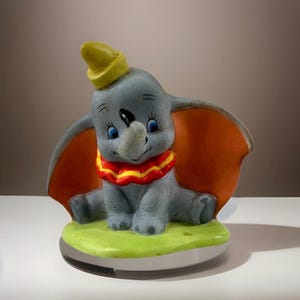 May include: A ceramic figurine of Dumbo the elephant, featuring a gray body, large orange ears, and a yellow hat. The figurine has a red and yellow ruffled collar and sits on a green base. The base is approximately 4 inches wide.