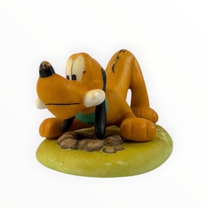 Pode incluir: Uma estatueta vintage do Pluto numa pose brincalhona. O cão laranja tem um nariz e orelhas pretas, com uma coleira verde. A estatueta está numa base verde-amarela com uma mancha castanha.