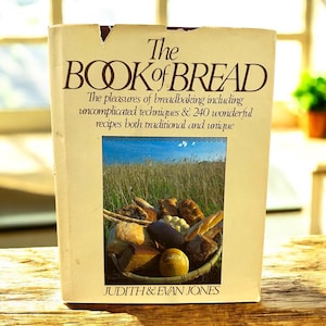 Puede incluir: Un libro de cocina vintage titulado "The Book of Bread" con una portada de color crema. La portada presenta texto y una imagen de varios panes en una cesta. El libro promete técnicas sencillas y 240 recetas. Autores: Judith & Evan Jones.