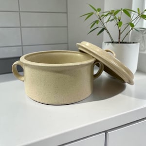 Puede incluir: Un cuenco de sopa de cerámica beige con una tapa a juego, ligeramente entreabierta. El cuenco tiene dos asas y una textura moteada. La tapa está apoyada en el lateral, revelando el interior. El cuenco está sobre una superficie blanca.