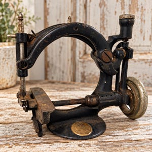 May include: A vintage black cast iron sewing machine with a curved arm, a metallic hand wheel, and a needle bar. The base features a circular brass emblem with embossed details. The metal surfaces show signs of age, oxidation, and wear throughout.