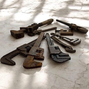 May include: A collection of antique wrenches and tools, in various sizes and styles, arranged on a light-colored surface. The tools show signs of age and wear, with a rusty, aged appearance. The collection includes adjustable wrenches and other hand tools.
