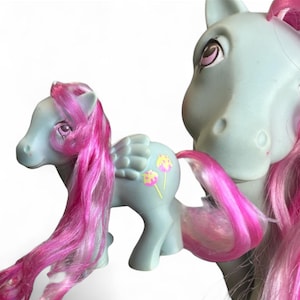 May include: Two vintage toy ponies. One is a light blue pony with pink hair and wings, and yellow and pink flower designs on its flank. The other pony is a light blue pony with pink hair.