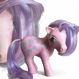 May include: A vintage pink toy pony with blue eyes and a lavender mane and tail. The pony's body is decorated with blue and pink flower designs. The image also shows a partial view of a second pony with lavender hair.