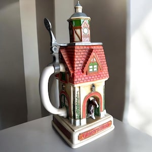 Può includere: Un boccale da birra decorativo in ceramica a forma di edificio con tetto di tegole rosse e una piccola torre. Il boccale ha un manico bianco e un coperchio incernierato in metallo. La base recita "Grant's Farm".