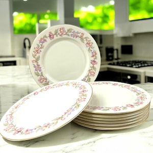 May include: A set of white ceramic plates with a floral design in pink, purple, and green. The plates have a gold rim and are stacked on a marble surface. The floral pattern encircles the plate's edge, creating an elegant aesthetic. The image shows a stack of plates and two individual plates.