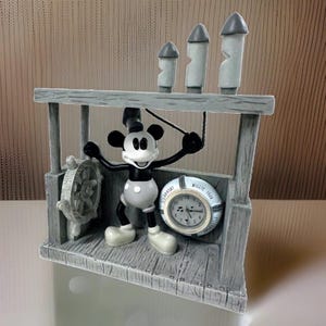 May include: A grey-toned decorative clock featuring a black and white Mickey Mouse figure standing on a wooden-style deck. The figure holds a rope near a ship's wheel. A round clock face is inscribed with the text Steamboat Willie 1928.