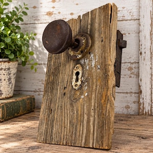 May include: A weathered, rectangular piece of reclaimed wood featuring a dark, rusted metal round doorknob and a matching metal keyhole plate. The wood displays a natural grain with visible cracks and remnants of white paint on the surface.