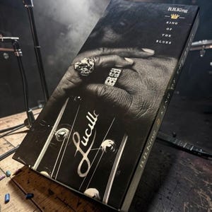 Könnte beinhalten: Schwarz-weiß Box-Set von B.B. King, betitelt "King of the Blues". Das Bild zeigt eine Hand mit Ringen, die auf einer Gitarrenkopfplatte mit dem Namen "Lucille" ruht. Das Box-Set befindet sich auf einer Holzoberfläche.