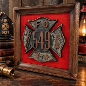 May include: A square wooden frame containing a red felt background and a metallic Maltese cross emblem. The emblem features the letters FD, the number 549, and relief icons of a ladder, a fire hydrant, and crossed axes in a bronze-toned finish.