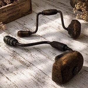 Op de afbeelding: Twee vintage handboren met donkere, verweerde metalen frames en versleten houten handvatten. Elk gereedschap heeft een zwengelmechanisme met een draaibare greep en een massieve houten knop aan het uiteinde, met een rustieke, verouderde textuur.