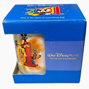 May include: A Walt Disney World 2001 mug in a blue box. The mug features Mickey Mouse, Goofy, and the number "01" in a red color. The box has the text "This is the start of something Big!"