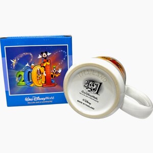 May include: White ceramic mug with a handle, featuring a colorful design of Disney characters and the number "2001". The mug is accompanied by a blue box with matching graphics. The bottom of the mug reads "Made in Thailand".