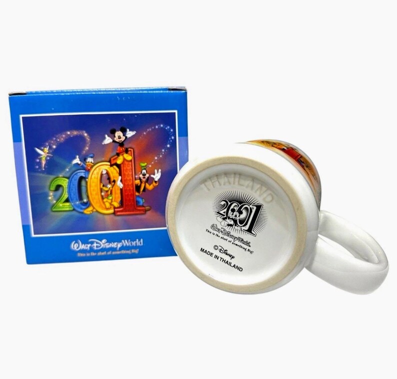 May include: White ceramic mug with a handle, featuring a colorful design of Disney characters and the number "2001". The mug is accompanied by a blue box with matching graphics. The bottom of the mug reads "Made in Thailand".