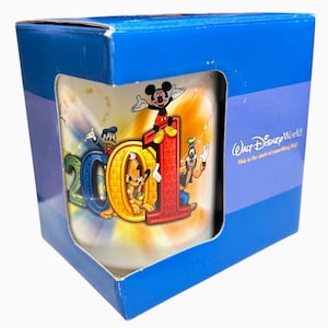 May include: A Walt Disney World 2001 mug in a blue box. The mug features Mickey Mouse, Donald Duck, Goofy, and Pluto with the numbers "2001". The box has the Walt Disney World logo.