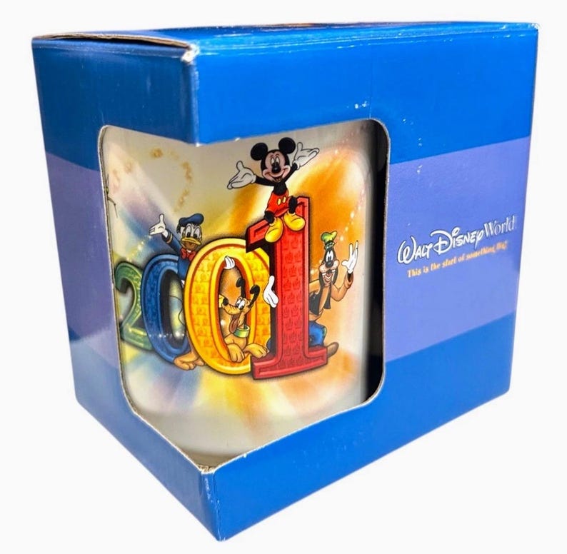 May include: A Walt Disney World 2001 mug in a blue box. The mug features Mickey Mouse, Donald Duck, Goofy, and Pluto with the numbers "2001". The box has the Walt Disney World logo.