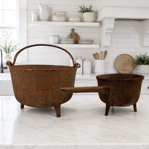 May include: Two rusted cast iron vessels with a dark brown, textured finish. The larger vessel is a round pot with three short legs and a curved metal handle. The smaller vessel is a round pot with three short legs and a single straight handle.