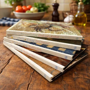 Puede incluir: Una pila de libros de cocina antiguos con títulos como "American Cooking: Creole and Acadian" y "American Cooking: New England". Los libros tienen cubiertas coloridas y están colocados sobre una superficie de madera, lo que sugiere un tema culinario.