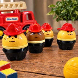Op de afbeelding: Vier kleine plastic speelgoedfiguren in de vorm van ronde karakters met felrode brandweerhelmen. Elk figuurtje heeft een geel of bruin hoofd met een eenvoudig geschilderd gezicht en een zwarte basis die een uniform voorstelt. Een rode speelgoedbrandweerwagen is zichtbaar.